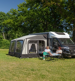Een camper met een grote voortent staat op een groene campingplaats. Twee mensen zitten ontspannen in stoelen voor de tent, met bomen en blauwe lucht op de achtergrond.